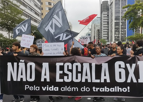 Manifestantes em uma avenida urbana seguram uma grande faixa preta com letras brancas onde se lê: "NÃO À ESCALA 6X1 - Pela vida além do trabalho!". Ao fundo, bandeiras e prédios compõem o cenário do protesto por melhores condições laborais.