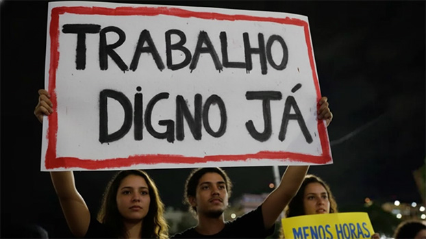 Jovens em uma manifestação noturna segurando cartazes artesanais. O cartaz central, branco com bordas vermelhas, diz "TRABALHO DIGNO JÁ". Ao lado, um cartaz amarelo traz a frase "MENOS HORAS MAIS VIDA".